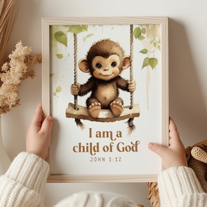 May include: A framed print featuring a watercolour illustration of a monkey sitting on a swing. The monkey is brown with a light brown belly and has a big smile. The background is a light green and white watercolour wash. The text below the monkey reads "I am a child of God" and "John 1:12".