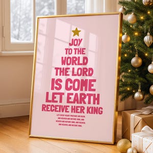 Puede incluir: Un estampado rosa enmarcado con borde dorado, con el texto "JOY TO THE WORLD THE LORD IS COME LET EARTH RECEIVE HER KING" en forma de árbol de Navidad. Una estrella dorada está en la parte superior. El estampado está apoyado contra una pared cerca de un árbol de Navidad.