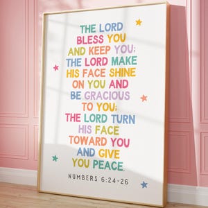 Puede incluir: Un póster blanco con marco dorado presenta una superposición de texto colorido de un versículo bíblico de Números 6:24-26. El texto dice: "Que el Señor te bendiga y te guarde; que el Señor haga brillar su rostro sobre ti y te sea propicio; que el Señor vuelva su rostro hacia ti y te dé paz." El versículo está impreso en un arcoíris de colores con estrellas esparcidas por todo el diseño.