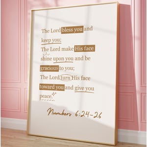 May include: A framed print with a gold frame featuring a religious quote from Numbers 6:24-26 in a handwritten style. The text is in a dark brown color against a cream background. The print is displayed against a pink wall.