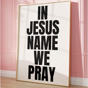 Puede incluir: Un cuadro enmarcado con las palabras "IN JESUS NAME WE PRAY" en letras negras en negrita sobre un fondo blanco. El marco es dorado y la obra de arte está apoyada contra una pared rosa.