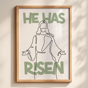 Puede incluir: Una impresión artística enmarcada con un dibujo lineal minimalista de una figura con los brazos extendidos. Las palabras "HE HAS RISEN" están en letras verdes en bloque encima y debajo de la figura. La obra de arte está sobre un fondo blanco.