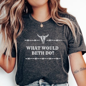 Peut inclure: Un t-shirt gris foncé avec un graphique blanc d'un crâne de taureau et de barbelés. Le texte "What Would Beth Do?" est imprimé sous le graphique.