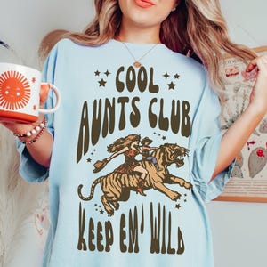 May include: A light blue t-shirt with a brown graphic of a woman riding a tiger. The graphic includes the text "Cool Aunts Club Keep 'Em Wild".