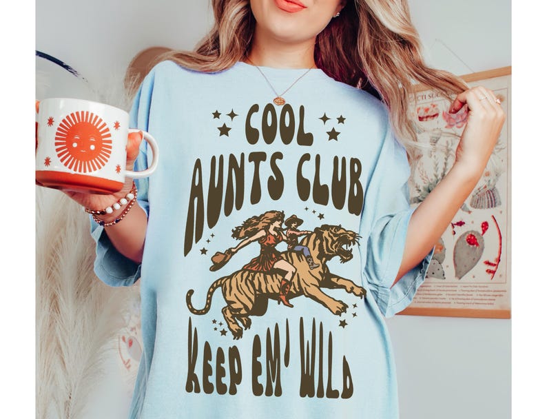 May include: A light blue t-shirt with a brown graphic of a woman riding a tiger. The graphic includes the text "Cool Aunts Club Keep 'Em Wild".