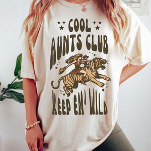 May include: A white t-shirt with the text "Cool Aunts Club Keep 'em Wild" in brown lettering. The graphic features a woman riding a tiger.