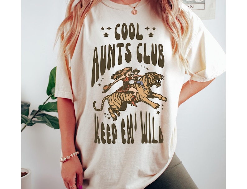 May include: A white t-shirt with the text "Cool Aunts Club Keep 'em Wild" in brown lettering. The graphic features a woman riding a tiger.
