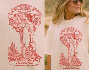 May include: A cream-colored t-shirt with a red illustration of a woman standing in a tree with apples. The text "The Best Gals" is on a banner above the woman and the text "Read Smut" is on a banner below the woman.