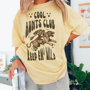 May include: A yellow t-shirt with the text "Cool Aunts Club Keep 'em Wild" in brown lettering. A cartoon image of a woman riding a tiger is above the text.