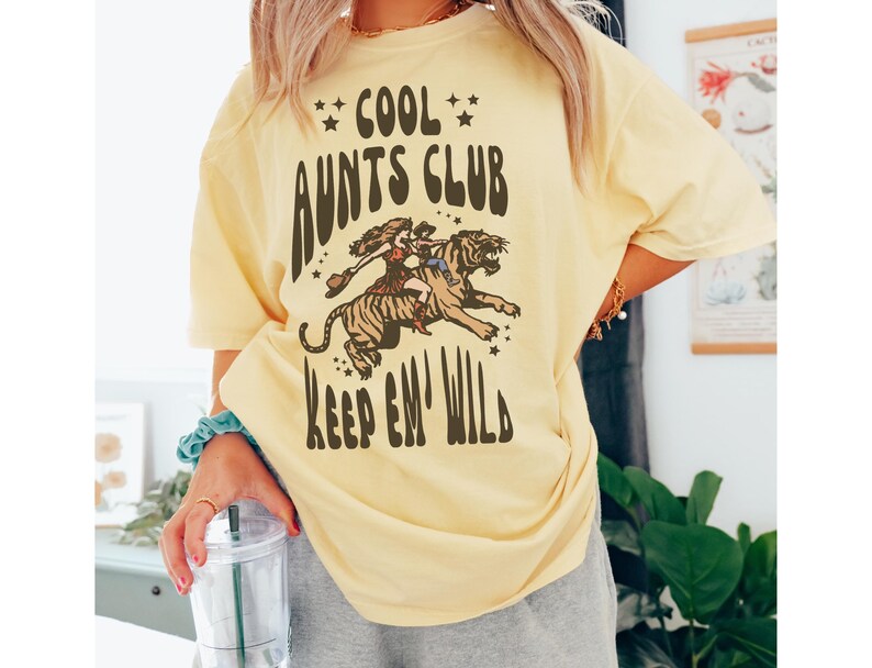 May include: A yellow t-shirt with the text "Cool Aunts Club Keep 'em Wild" in brown lettering. A cartoon image of a woman riding a tiger is above the text.