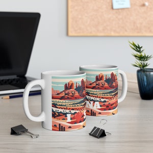 May include: Two white ceramic mugs with a colorful illustration of a desert landscape and the text "I Love Sedona" on one mug. The mugs are on a wooden table.