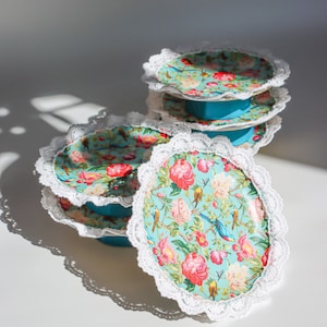 May include: A set of four turquoise cake stands with white lace trim and floral patterns. The stands are stacked on top of each other, with the largest stand at the bottom and the smallest stand at the top.