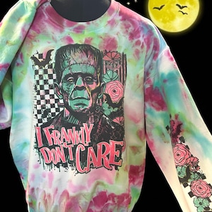 Könnte beinhalten: Ein Sweatshirt mit Batikmuster, das ein Frankenstein-Motiv und den Text "I Frankly Don't Care" zeigt. Die langen Ärmel haben ein Blumenmuster. Die Farben sind Pink, Grün und Weiß.