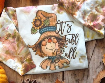 Könnte beinhalten: Ein weißes Sweatshirt mit einem herbstlichen Grafikmotiv einer Vogelscheuche mit Hut und Sonnenblume. Der Text "It's Fall Y'all" ist darunter gedruckt. Die Ärmel haben ein Batikmuster in Braun, Rosa und Gelb.