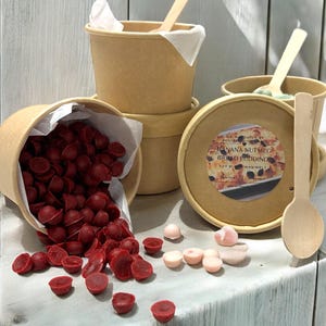 May include: A selection of soy wax melts in shades of red and pink, presented in a brown paper cup. The image also features several brown paper cups, wooden spoons, and a lid with the text "Banana Nutmeg Bread Pudding Wax Melt".