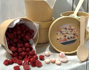 May include: A display of soy wax melts in various colors and shapes, including red and pink, spilling from a brown paper cup. The image also shows several brown paper cups, wooden spoons, and a lid with text that reads "Banana Nutmeg Bread Pudding Wax Melt."