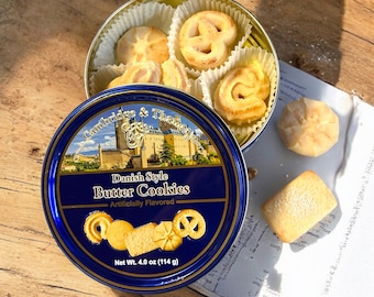 May include: A blue tin with a gold trim and a picture of a castle on it. The tin contains Danish style butter cookies. The text on the tin reads "Cambridge & Thames" and "Danish Style Butter Cookies - Artificially Flavored - Net Wt. 4.0 oz (114 g)".