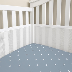 May include: A baby crib with a white frame and a blue fitted sheet. The sheet is patterned with white crescent moons. The crib has a white mesh lining and vertical white bars.