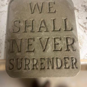 Könnte beinhalten: Rechteckige, hellgraue Betonplatte mit den Worten "WE SHALL NEVER SURRENDER" in die Oberfläche gemeißelt. Der Text ist in einer fetten, serifenlosen Schrift.