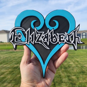 May include: A heart-shaped sign featuring a bright cyan outline and a black center. The name Elizabeth is written across the center in a stylized, sharp white font with black outlines, creating a layered, three-dimensional appearance.