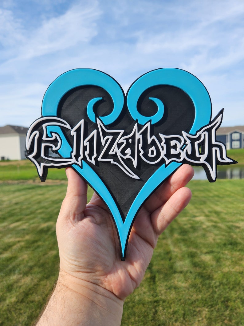 May include: A heart-shaped sign featuring a bright cyan outline and a black center. The name Elizabeth is written across the center in a stylized, sharp white font with black outlines, creating a layered, three-dimensional appearance.