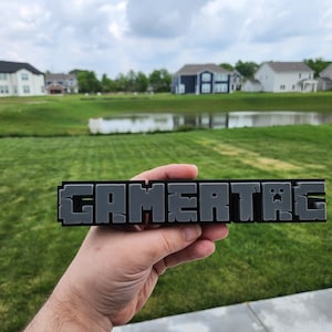 May include: A black and grey 3D printed sign that reads "GAMERTAG" in a pixelated font. The sign is being held in front of a green grassy lawn with a pond and houses in the background.
