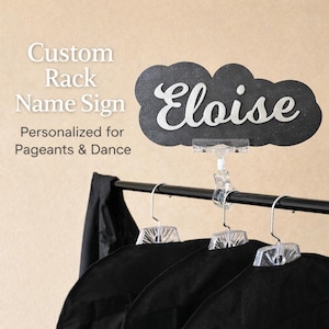 May include: A custom rack name sign with the name "Eloise" in white script on a black cloud-shaped background. The sign is personalized for pageants and dance. Black garment bags hang on a rack.