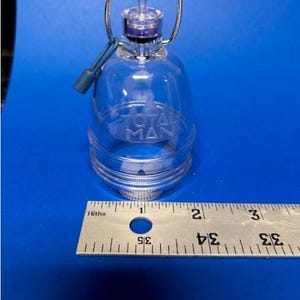 May include: A clear, bell-shaped container with a silver handle and a blue tether. The container has the text "TOTAL MAN" embossed on the front. A ruler is placed next to the container, with measurements in inches.