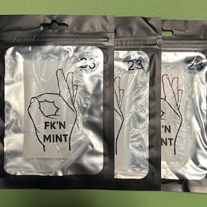 May include: Three sealed, clear plastic packages with a black border. Each package features a white graphic of a hand gesture and the words "FK'N MINT" in black. A circular sticker with the number "23" is also visible on each package.