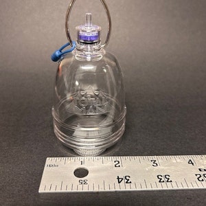 May include: A clear plastic bottle with a silver ring handle and a blue rubber seal. The bottle has a clear plastic cap with a purple and black top. The bottle is sitting on a ruler that is marked in inches.
