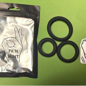 May include: Black rubber rings in varying sizes, a black plastic bag with a hand gesture graphic and the text "F*K'N MINT", and a sticker with the same graphic and text. The items are on a green surface.