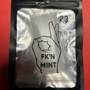 以下が含まれることがあります： 黒いビニール袋に入った透明な長方形のアイテム。アイテムには、手の「OK」ジェスチャーの白黒グラフィックがあり、その下に「FK'N MINT」という言葉があります。バッグには数字の「23」が印刷されています。