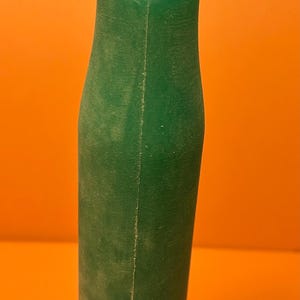 Puede incluir: Un objeto cilíndrico de color verde oscuro translúcido con una parte superior inclinada y abierta. El artículo presenta una costura vertical visible a lo largo de su longitud, lo que indica una construcción moldeada. La superficie es mate.