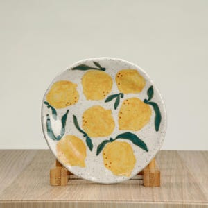 May include: A white ceramic plate with a speckled glaze features a hand-painted design of yellow lemons with green leaves. The plate is displayed on a wooden stand.