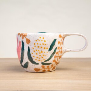 May include: White ceramic mug with a speckled glaze and a floral design in shades of pink, green, and brown. The mug has a rounded shape and a handle.