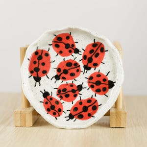 May include: A round, white ceramic dish with a wavy, scalloped edge, featuring a hand-painted pattern of seven bright red ladybirds with black spots scattered across the surface. The ceramic has a speckled texture throughout the white base.