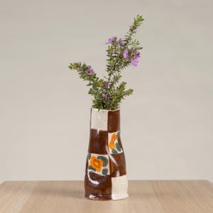 May include: A handcrafted ceramic vase featuring a brown and white chequered design, decorated with orange floral motifs. The vase contains a display of green stems and small purple flowers, set against a plain backdrop.