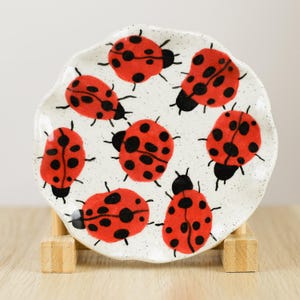May include: A small, white ceramic plate with a scalloped edge, decorated with red ladybug illustrations with black spots. The plate is supported by a wooden stand and has a speckled background.