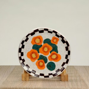 May include: A white ceramic plate with a black and brown chequered border. The centre of the plate features a floral design with orange and yellow flowers and green leaves.