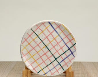 May include: A small white ceramic plate with a pink, yellow, green, and blue checkered pattern. The plate is on a wooden stand.