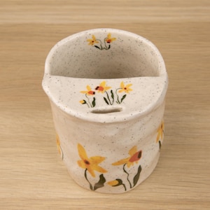 May include: A speckled off-white ceramic wall pocket featuring hand-painted yellow daffodil flowers with green stems and leaves. The vessel has a semi-circular opening at the top and a small horizontal slot below the rim for mounting purposes.