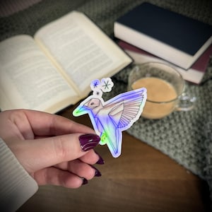 May include: A holographic sticker of a geometric hummingbird with a colorful, iridescent finish. The sticker has a white outline and is held in front of a blurred background with books and a cup of tea.