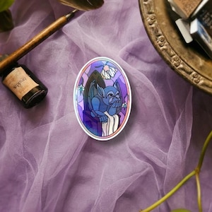 May include: An oval sticker featuring a blue gargoyle sitting on a stone ledge within a purple and pink stained glass window frame. The design includes a small white moth hovering above the gargoyle, with intricate dark outlines and soft shading.