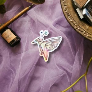 May include: A sticker featuring an origami-style bird with iridescent, multi-colored geometric panels and wings spread in flight. Above the bird are three small, colorful star-shaped icons in blue, green, and black, all set against a light purple fabric.