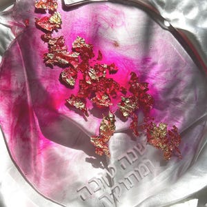May include: A pomegranate-shaped decorative tray with a translucent pink and white marbled design. Gold leaf accents are scattered across the surface. The tray has etched Hebrew text. The tray is on a white surface.