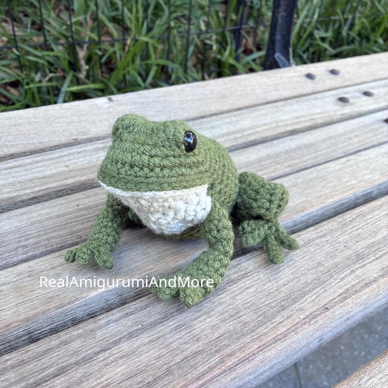 May include: A crocheted frog amigurumi toy featuring a textured olive green body, a cream-colored underbelly, and two small black plastic eyes. The text RealAmigurumiAndMore is printed in white on the lower left side of the image.