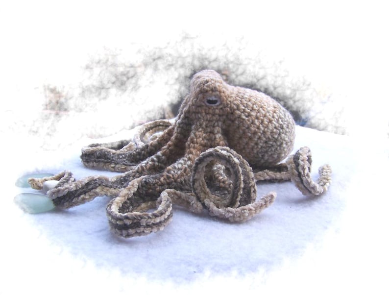May include: A crocheted octopus with brown and white yarn. The octopus is lying on a white surface.