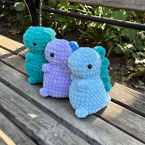 Puede incluir: Tres peluches de dinosaurio hechos a mano con ganchillo en tonos turquesa, morado y azul. Cada dinosaurio tiene un cuerpo texturizado, brazos y piernas pequeños y una espina dorsal de color contrastante. Los juguetes se exhiben en un banco de madera.