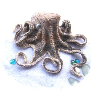 May include: A crocheted octopus with brown and white yarn. The octopus has eight tentacles and is sitting on a white surface with blue and green glass beads.