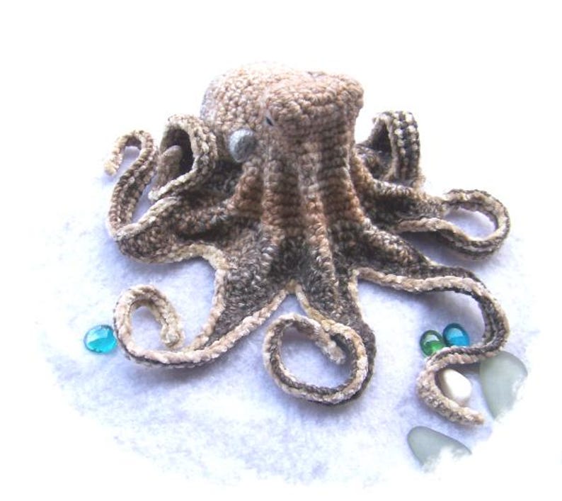 May include: A crocheted octopus with brown and white yarn. The octopus has eight tentacles and is sitting on a white surface with blue and green glass beads.